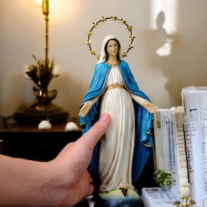 Op de afbeelding: Een standbeeld van de Maagd Maria, met een witte jurk en een blauwe mantel. Het beeld heeft een gouden halo met sterren en staat op een donkere basis. De afbeelding is van een religieuze figuur.