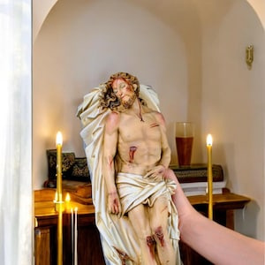 Peut inclure: Statue religieuse repr&eacute;sentant une figure bless&eacute;e, drap&eacute;e dans un tissu. La figure a une barbe et de longs cheveux, avec une couronne d'&eacute;pines &agrave; la base. La statue est tenue devant un meuble en bois avec des bougies allum&eacute;es.