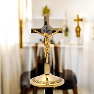 May include: A gold-coloured crucifix featuring a detailed figure of Jesus Christ. The cross has a decorative emblem at the intersection and stands on a round, ornate base. Another cross is visible in the background.