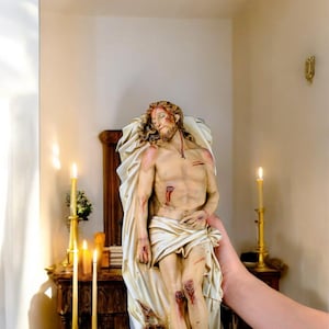 Peut inclure: Statue d&eacute;taill&eacute;e d'une figure bless&eacute;e, drap&eacute;e dans un tissu blanc. La figure a une barbe et de longs cheveux, avec une couronne d'&eacute;pines &agrave; la base. La statue est tenue devant un autel en bois avec des bougies allum&eacute;es.