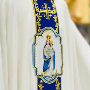 May include: White religious vestment with blue and gold embroidered details. The vestment features a blue collar and a central panel with a gold cross design and an embroidered image of the Virgin Mary and child.