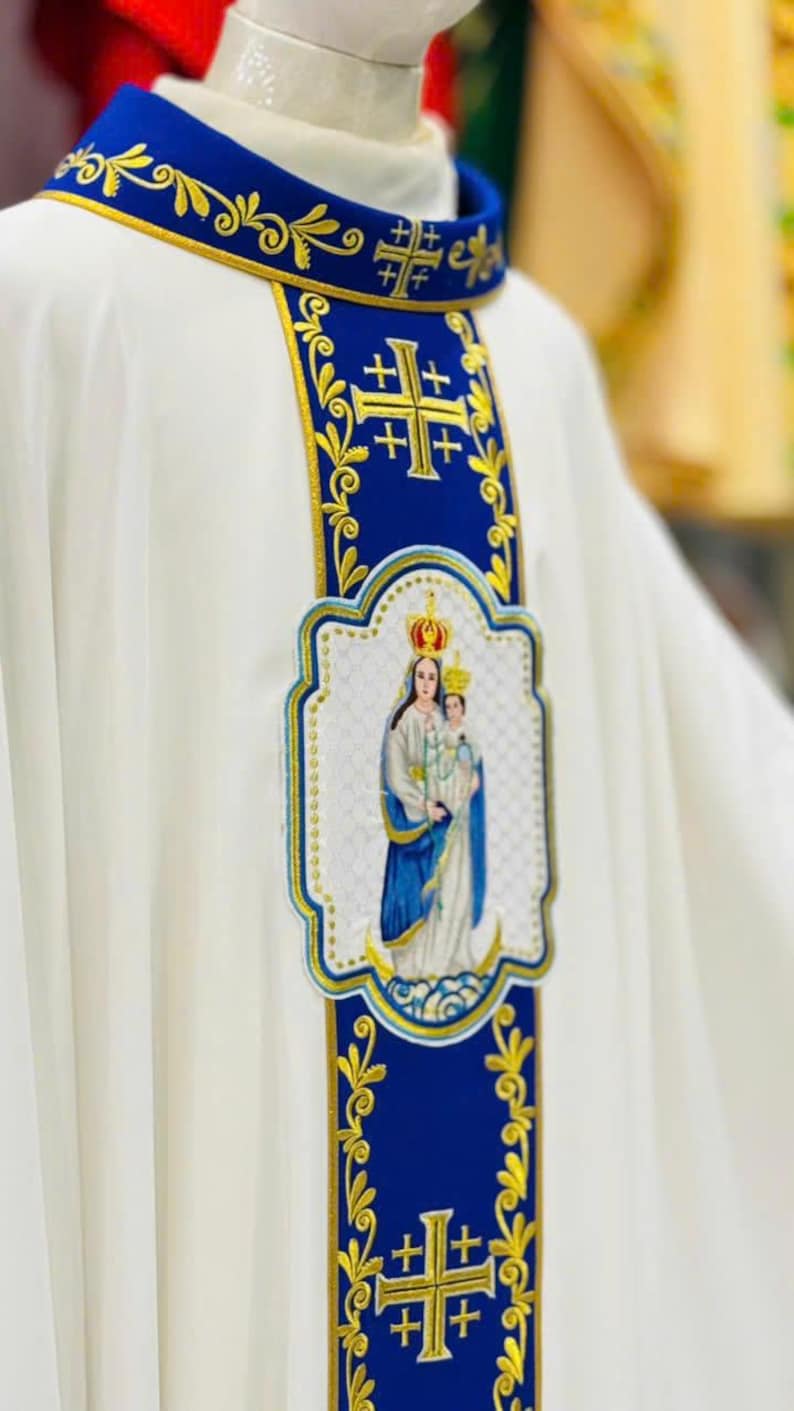 May include: White religious vestment with blue and gold embroidered details. The vestment features a blue collar and a central panel with a gold cross design and an embroidered image of the Virgin Mary and child.