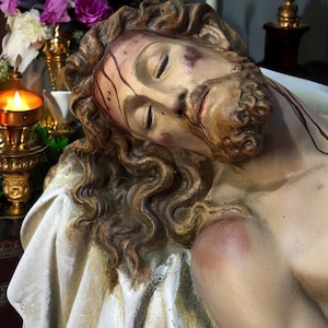 Peut inclure: Une sculpture d&eacute;taill&eacute;e repr&eacute;sentant une figure aux yeux ferm&eacute;s, aux cheveux bruns et &agrave; la barbe. Le visage de la figure montre des signes de blessures, avec des stries et des marques rouges. La sculpture repose sur un tissu blanc, avec un arri&egrave;re-plan flou de fleurs et de bougies.