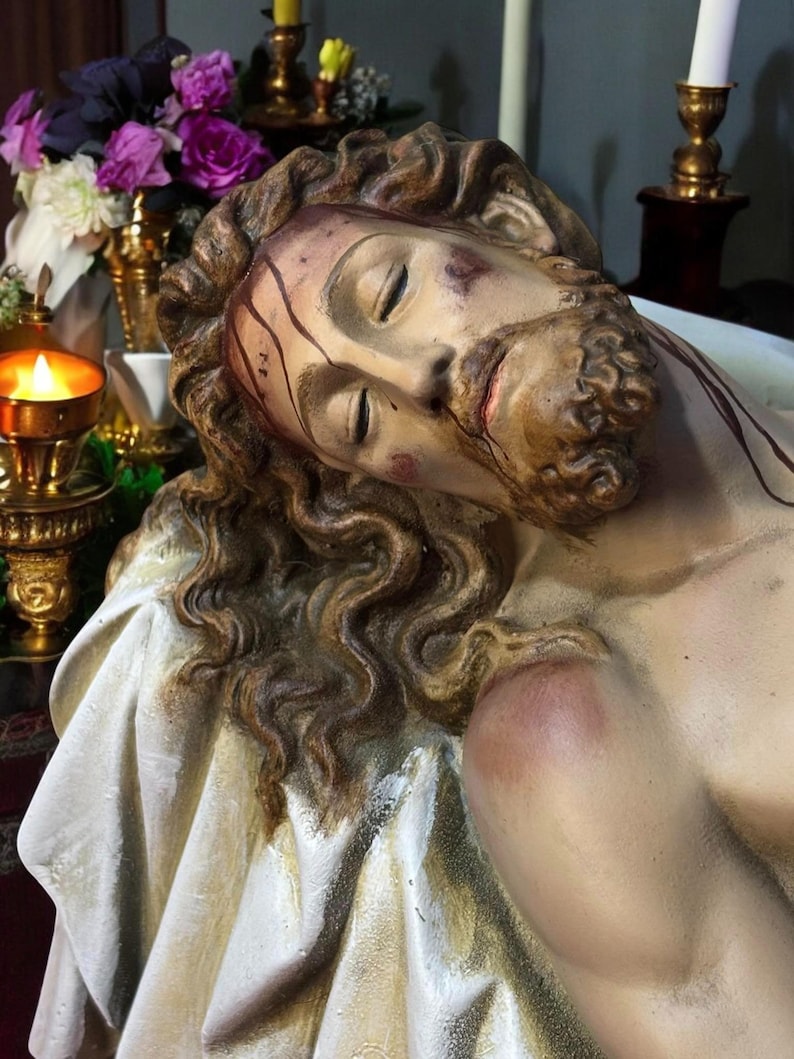 Peut inclure: Une sculpture d&eacute;taill&eacute;e repr&eacute;sentant une figure aux yeux ferm&eacute;s, aux cheveux bruns et &agrave; la barbe. Le visage de la figure montre des signes de blessures, avec des stries et des marques rouges. La sculpture repose sur un tissu blanc, avec un arri&egrave;re-plan flou de fleurs et de bougies.