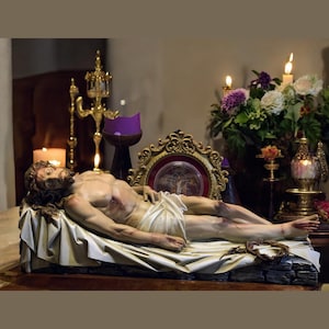 Peut inclure: Une sculpture d&eacute;taill&eacute;e d'un homme allong&eacute;, drap&eacute; dans un tissu blanc, avec une couronne d'&eacute;pines &agrave; proximit&eacute;. La sc&egrave;ne est &eacute;clair&eacute;e par des bougies et une composition florale, cr&eacute;ant une atmosph&egrave;re de recueillement. Une image encadr&eacute;e en or est en arri&egrave;re-plan.