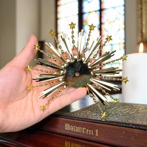 Könnte beinhalten: Ein kreisförmiges Zierobjekt mit einer zentralen dunklen Steinscheibe, umgeben von strahlenförmigen, goldfarbenen Metallspitzen und kleinen goldenen Sternen an dünnen Drahtstielen. Das Stück hat ein Loch in der Mitte und ein Strahlenkranz-Design.