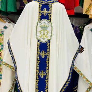 May include: White liturgical vestment with blue and gold embroidered details. The vestment features a blue collar and vertical blue panels with gold crosses. A decorative emblem is centered on the back.