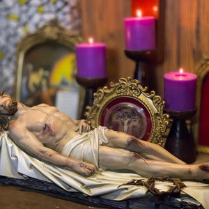 Peut inclure: Une sculpture d&eacute;taill&eacute;e d'une figure couch&eacute;e, repr&eacute;sentant un homme bless&eacute;, drap&eacute; dans un tissu blanc. La figure est entour&eacute;e de bougies violettes allum&eacute;es et de cadres dor&eacute;s orn&eacute;s, sugg&eacute;rant un th&egrave;me religieux. Une couronne d'&eacute;pines repose pr&egrave;s des pieds.