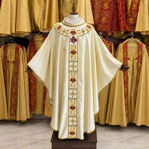 Op de afbeelding: Een crèmekleurig liturgisch gewaad met gouden en rode geborduurde details. Het gewaad wordt op een mannequin getoond, met andere soortgelijke kledingstukken op de achtergrond. Het ontwerp omvat een kruis en bloemmotieven.