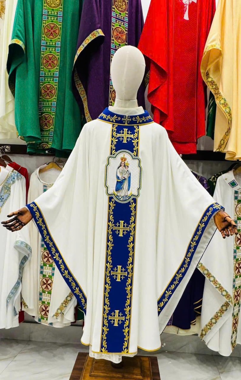 May include: White liturgical vestment with blue and gold embroidered details. The vestment features a central panel with an image of the Virgin Mary and Jesus. Other vestments in green, purple, and red are visible in the background.