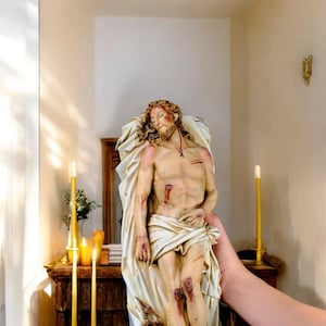 Peut inclure: Statue d&eacute;taill&eacute;e repr&eacute;sentant une figure bless&eacute;e, drap&eacute;e de tissu. La figure a une barbe et de longs cheveux, avec des blessures visibles sur le torse et les jambes. La statue est tenue devant un meuble en bois avec des bougies.