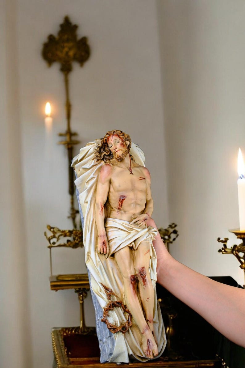 Peut inclure: Une figurine religieuse d&eacute;taill&eacute;e repr&eacute;sentant une figure avec des blessures, drap&eacute;e dans un tissu. La figure porte une couronne d'&eacute;pines et est tenue par une personne. L'arri&egrave;re-plan comprend une bougie et des ornements en m&eacute;tal.