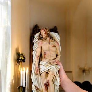 Peut inclure: Statue d&eacute;taill&eacute;e repr&eacute;sentant une figure avec des blessures, drap&eacute;e dans un tissu blanc. La figure a de longs cheveux et une barbe, avec des blessures visibles. La statue est tenue par une personne, avec des bougies et un support d&eacute;coratif en arri&egrave;re-plan.
