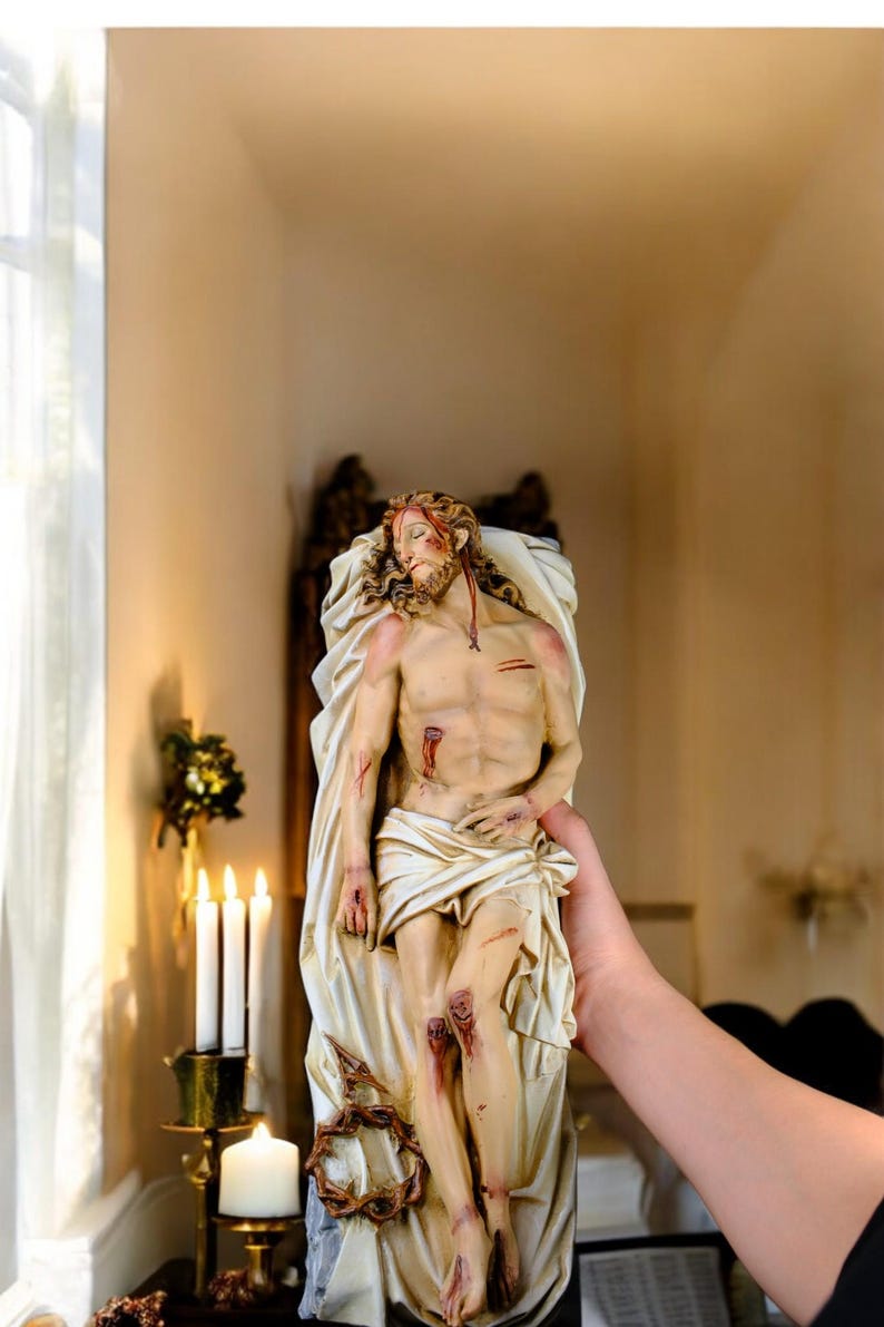 Peut inclure: Statue d&eacute;taill&eacute;e repr&eacute;sentant une figure avec des blessures, drap&eacute;e dans un tissu blanc. La figure a de longs cheveux et une barbe, avec des blessures visibles. La statue est tenue par une personne, avec des bougies et un support d&eacute;coratif en arri&egrave;re-plan.