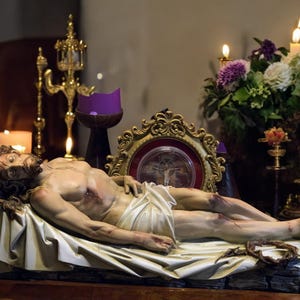 Peut inclure: Une sculpture d&eacute;taill&eacute;e d'un homme allong&eacute;, avec des blessures visibles et un drap blanc sur le bas du corps. La sculpture repose sur une surface en bois, entour&eacute;e de bougies, de fleurs et de d&eacute;corations dor&eacute;es orn&eacute;es, cr&eacute;ant une atmosph&egrave;re de r&eacute;v&eacute;rence.
