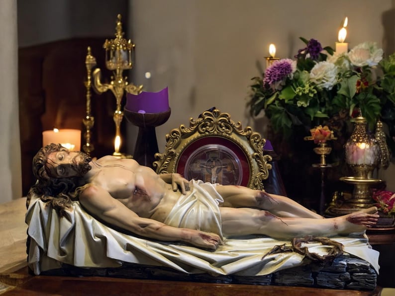 Peut inclure: Une sculpture d&eacute;taill&eacute;e d'un homme allong&eacute;, avec des blessures visibles et un drap blanc sur le bas du corps. La sculpture repose sur une surface en bois, entour&eacute;e de bougies, de fleurs et de d&eacute;corations dor&eacute;es orn&eacute;es, cr&eacute;ant une atmosph&egrave;re de r&eacute;v&eacute;rence.