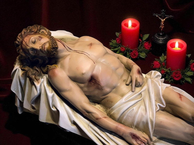 Peut inclure: Une sculpture d'un homme allong&eacute;, avec des blessures visibles sur son corps. Deux bougies rouges avec des flammes allum&eacute;es flanquent la sculpture, ainsi qu'un petit crucifix. Le fond est rouge fonc&eacute;.