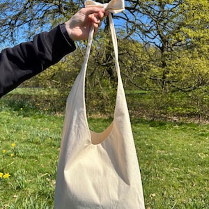 K&ouml;nnte beinhalten: Eine schlichte wei&szlig;e Canvas-Tragetasche mit einem langen Riemen, der zu einer Schleife gebunden ist. Die Tasche wird in einer Hand vor einem gr&uuml;nen grasbewachsenen Hintergrund gehalten.