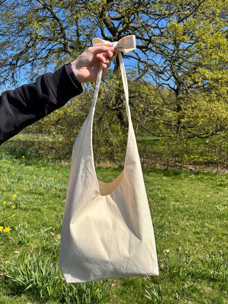K&ouml;nnte beinhalten: Eine schlichte wei&szlig;e Canvas-Tragetasche mit einem langen Riemen, der zu einer Schleife gebunden ist. Die Tasche wird in einer Hand vor einem gr&uuml;nen grasbewachsenen Hintergrund gehalten.