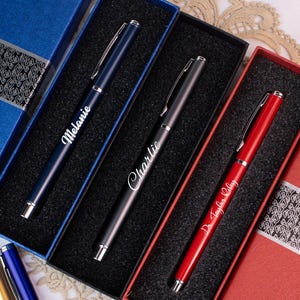 May include: A collection of personalized pens in various colors, including blue, black, and red, each displayed in a matching gift box. The pens are engraved with names like "Melanie" and "Charlie". The boxes are blue and red with decorative silver accents.
