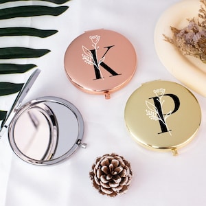 May include: Three personalized compact mirrors in rose gold and gold, each with a floral design and a letter, including "K" and "P". One mirror is open, showing a reflective surface. A pinecone and a decorative tray with dried flowers are also visible.