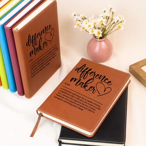 May include: A collection of notebooks with leather covers in various colours, including brown and black. The brown notebook features the words "difference maker" and a heart symbol. A definition of the term is also printed.