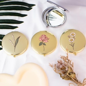 May include: A collection of gold-colored compact mirrors with floral designs. One mirror is open, revealing a reflective surface. The closed mirrors feature illustrations of a rose, daisies, and lily of the valley. The mirrors are round and have a shiny finish.