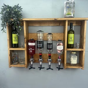 Op de afbeelding: Een rustieke houten barplank met meerdere compartimenten. Het bevat flessen drank, borrelglaasjes en een decoratieve lantaarn. De plank heeft ook vier drankdispensers en een kleine plant.