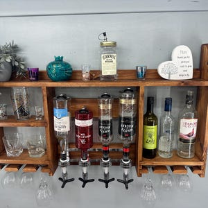 May include: A wooden bar shelf with multiple shelves and glass holders. It features bottles, shot glasses, and a decorative plant. The shelf includes liquor dispensers and various bottles of alcohol. The shelf is mounted on a light gray wall.