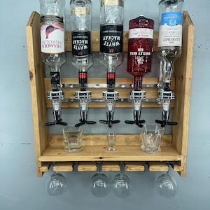 Op de afbeelding: Een houten drankdispenser met vijf omgekeerde flessen, elk met een ander etiket. Onder de flessen bevinden zich zilveren dispensers, een plank met borrelglaasjes en een rek voor wijnglazen. De dispenser is aan een grijze muur bevestigd.