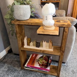 Pode incluir: Uma mesa lateral rústica de madeira com duas prateleiras. A prateleira superior contém uma planta em vaso e uma bola de lã branca em um suporte de madeira. A prateleira inferior exibe livros e uma vila de cerâmica.