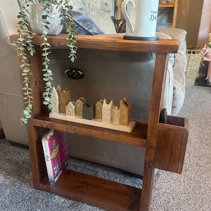 May include: A dark stained wooden side table with two shelves and a built-in cubby. A white mug with text sits on the top shelf. A small ceramic village and a book are on the lower shelf. A potted plant with trailing greenery is on the top shelf.