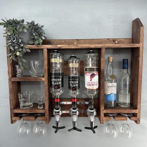 May include: A rustic, wooden bar shelf with three liquor dispensers, two shelves, and wine glass holders. The shelf holds bottles of liquor, shot glasses, and wine glasses. The dispensers are labeled "Whyte Mackay" and "Famous Grouse".