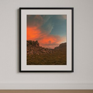May include: A scenic landscape photograph featuring a rocky mountain range with a vibrant orange and blue sunset sky. The sky is filled with dramatic clouds, creating a sense of depth and grandeur. The foreground shows a field of green grass and scattered rocks.