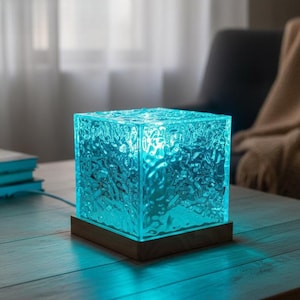 Peut inclure: Lampe cubique lumineuse avec un motif bleu glacé texturé. La lampe repose sur une base en bois et émet une lumière ambiante douce. La lampe est posée sur une table en bois, avec une plante et des livres en arrière-plan.