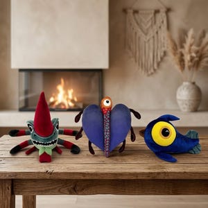 Puede incluir: Tres peluches con forma de criaturas caprichosas sobre una mesa de madera. Los juguetes incluyen una figura de insecto verde y rojo, una criatura azul en forma de corazón y un juguete azul con forma de pez con un gran ojo amarillo.