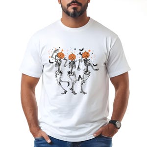 May include: White t-shirt featuring a Halloween design. The graphic depicts three dancing skeletons with pumpkin heads, surrounded by stars and bats. The skeletons are in black and white, and the pumpkins are orange.