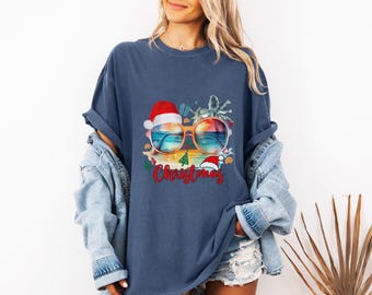 May include: Navy blue t-shirt with a graphic of sunglasses reflecting a beach scene, Santa hats, and the word "Christmas" in red. The design has a tropical holiday theme.