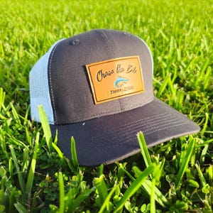 May include: A gray and white trucker hat with a brown leather patch that says "Chase the Bite" with a blue fish graphic.