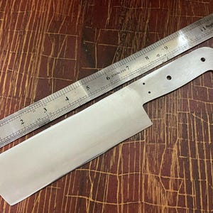 May include: A stainless steel knife blank with a ruler for scale. The blank is 12 inches long and has a rounded tip. The blank is unfinished and ready for grinding and polishing.