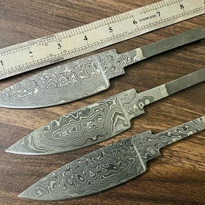 May include: Three Damascus steel knife blades with a wavy pattern. The blades are different lengths and have a polished finish.