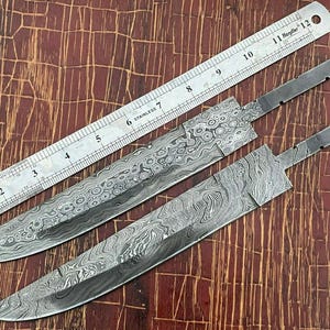 Puede incluir: Dos hojas de cuchillo de acero Damasco, una de 28 cm de largo y la otra de 30 cm de largo, se colocan sobre una superficie de madera con una regla para la escala.
