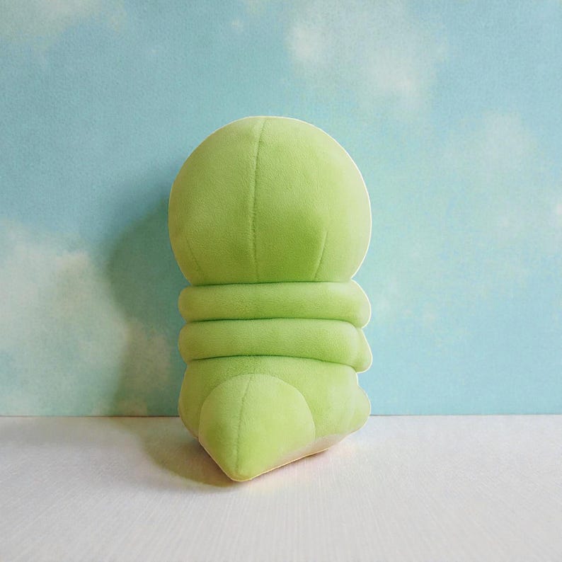 May include: A light green plush toy shaped like a caterpillar. The toy has a rounded head, segmented body, and a small, rounded tail. The plush is made of a soft, velvety material.