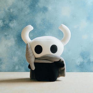 May include: A plush toy of a character with a white head, large black eyes, and curved horns. The character wears a gray scarf and a black body. The toy is set against a light blue background.