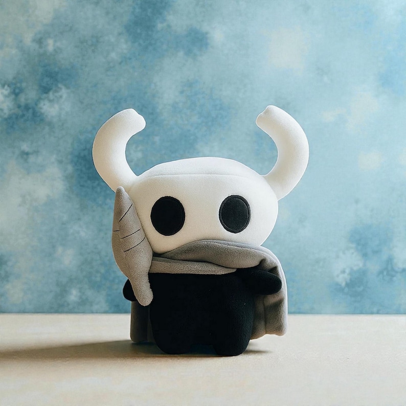 May include: A plush toy of a character with a white head, large black eyes, and curved horns. The character wears a gray scarf and a black body. The toy is set against a light blue background.