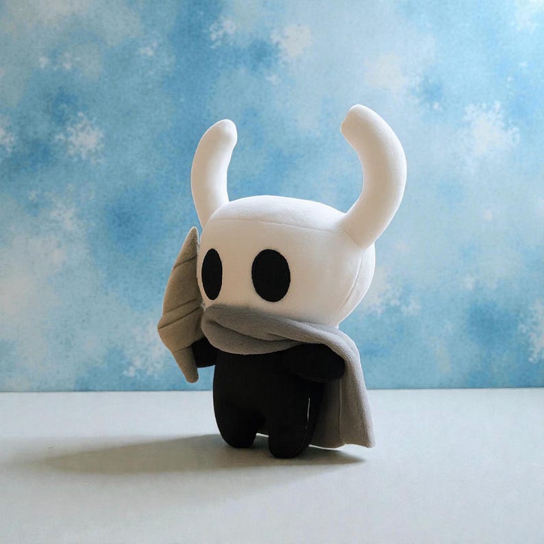 May include: A plush toy of a character from the video game Hollow Knight. The toy has a white head with curved horns, large black eyes, and a gray scarf and cape. The body is black, and it holds a gray weapon.