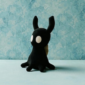 May include: A black plush toy with large white eyes and two tall, curved ears. The toy has a small, rounded body and four short, stubby legs. The background is a light blue with a mottled pattern.