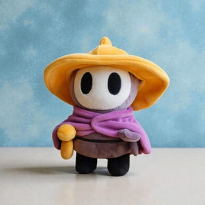 May include: A plush toy character with a large yellow hat, a white face with black oval eyes, and a purple scarf. The character has a brown body, black feet, and a small yellow mushroom-shaped accessory.