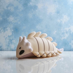 Pode incluir: Um brinquedo de pelúcia em forma de dinossauro bege claro com listras e detalhes em marrom escuro. O brinquedo tem uma cabeça branca com olhos pretos e uma parte inferior roxa clara. O fundo é um padrão de nuvens azul claro e branco.