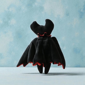 May include: A plush black bat-like figure with a red trim. The figure has a cape-like design with a ruffled red collar and two curved horns. The toy is set against a light blue background.